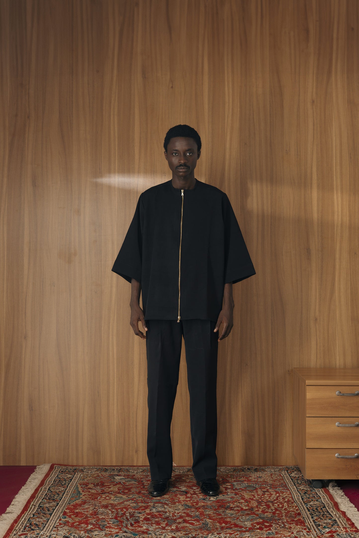 Model standing forward in a relaxed, minimalist black wide-sleeve shirt with a striking gold center zipper and matching black tailored trousers.